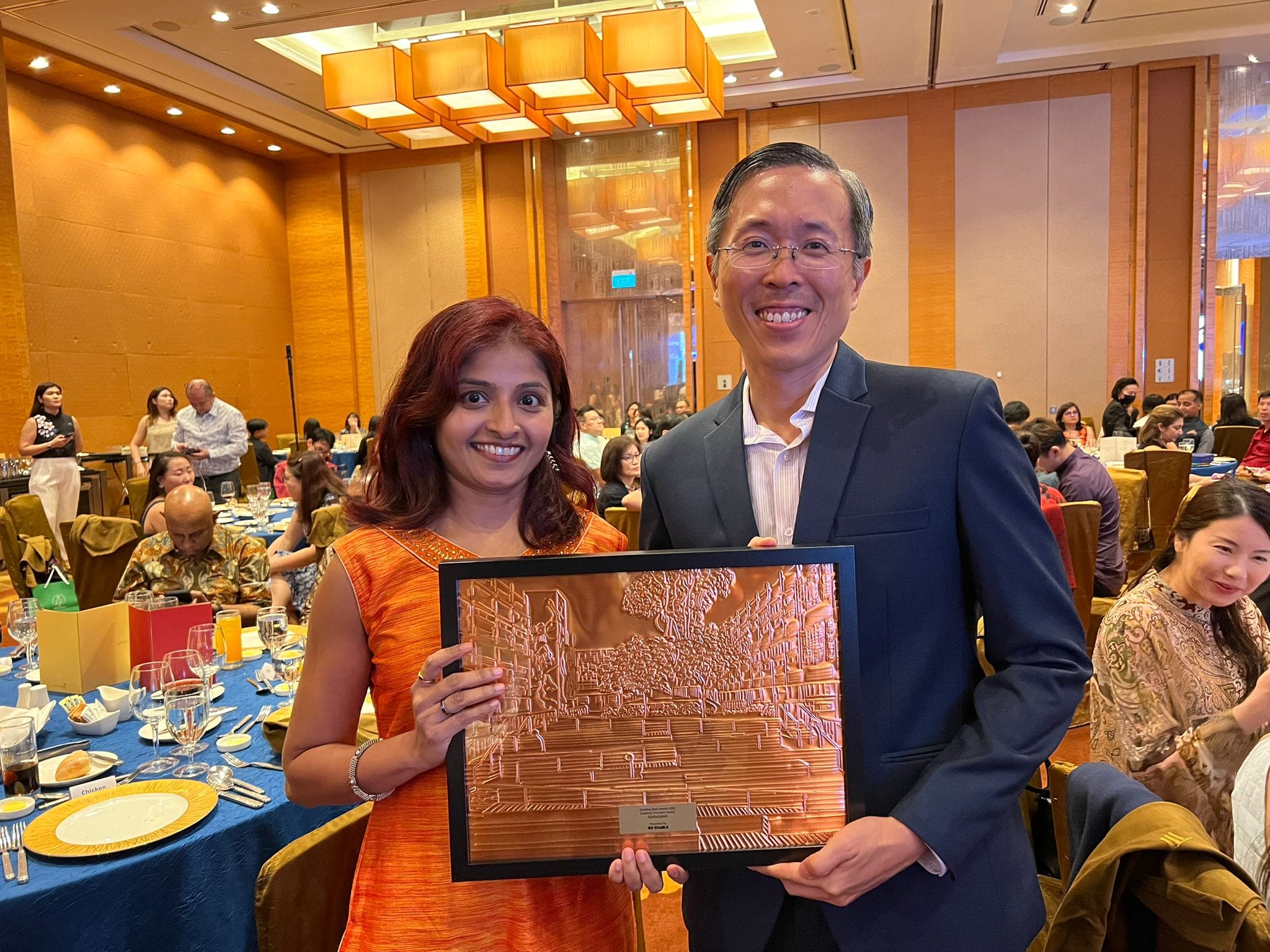 Two smiling people hold a framed copper relief plaque together at a formal banquet hall event.